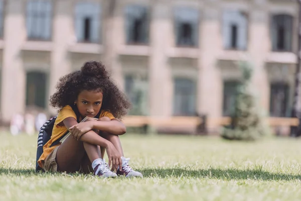 Upset african american schoolgirl looking at camera while sitting on lawn — Stock Photo