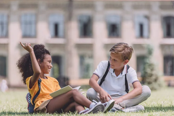 Alegre africano americano colegiala mostrando idea signo mientras sentado en el césped cerca multiétnico amigo - foto de stock