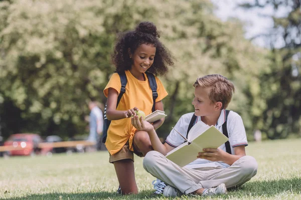 Cute schoolboy sitting on lawn and giving apple to smiling african american schoolgirl — Stock Photo
