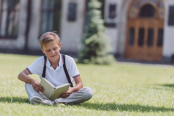 Entzückender Schuljunge sitzt auf dem Rasen in der Nähe der Schule und liest Buch — Stockfoto