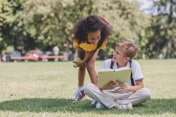 Sorridente scolaro africano americano in piedi vicino a un amico multiculturale seduto sul prato con libro — Foto stock