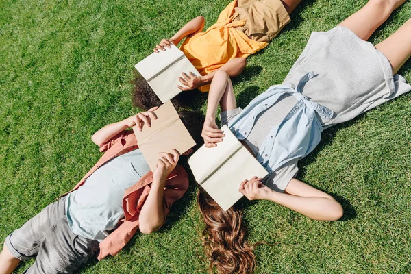 Top view of three multicultural schoolkids lying on lawn and covering faces with books — Stock Photo