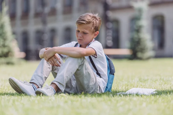 Pensive schoolboy with backpack sitting on green lawn near school — Stock Photo