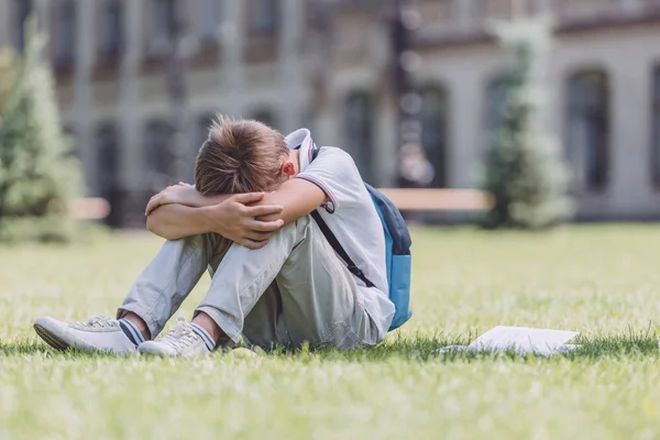 Estudante cansado com mochila sentado no gramado verde perto da escola — Fotografia de Stock