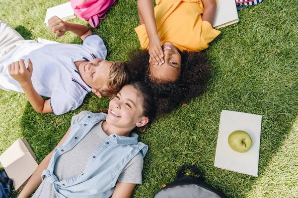 Blick von oben auf drei fröhliche multikulturelle Schulkinder, die lächelnd auf dem Rasen liegen — Stockfoto