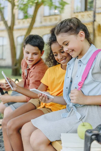 Tres escolares multiétnicos alegres usando teléfonos inteligentes mientras están sentados en el banco en el patio de la escuela - foto de stock