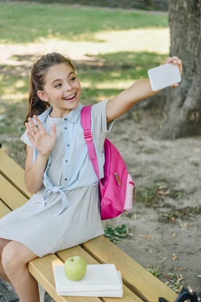 Colegiala feliz saludando de la mano mientras está sentado en el banco y tomando selfie con el teléfono inteligente - foto de stock