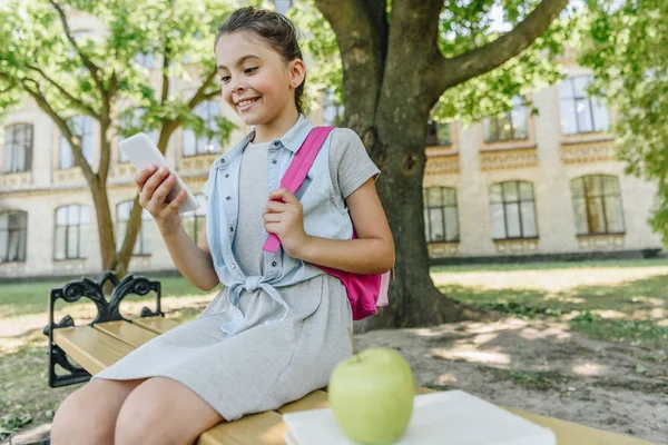 Colegiala alegre sentado en el banco en el parque y el uso de teléfono inteligente - foto de stock