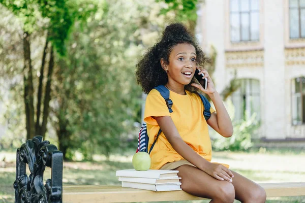 Sorprendió a la chica afroamericana hablando en un teléfono inteligente mientras estaba sentada en el banco cerca de libros y manzana - foto de stock