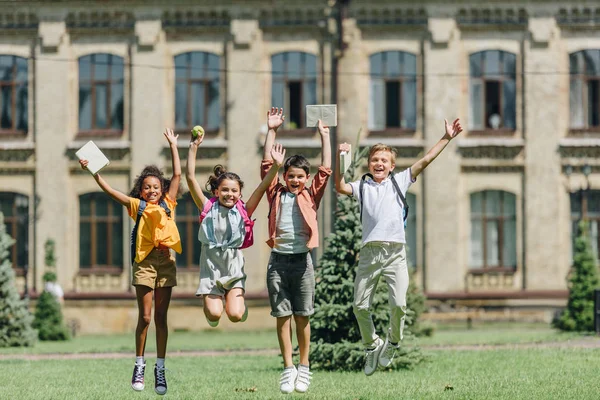 Quatro estudantes multiculturais felizes pulando enquanto segurando livros no gramado no parque — Fotografia de Stock