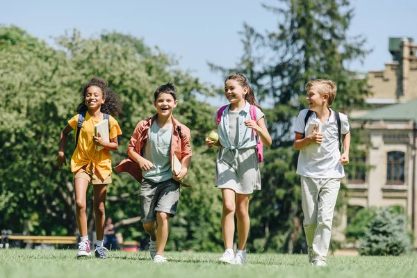 Quatro estudantes multiculturais felizes correndo no gramado no parque juntos — Fotografia de Stock