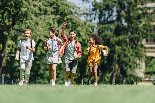 Cuatro escolares multiculturales felices corriendo en el césped verde en el parque - foto de stock