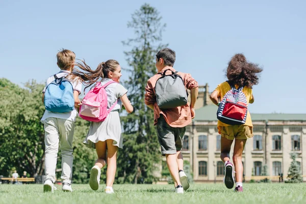 Visão traseira de quatro alunos multiétnicos com mochilas correndo no gramado no parque — Fotografia de Stock