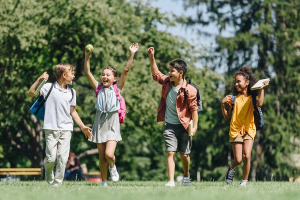 Quattro scolari eccitati che gesticolano con le mani alzate mentre corrono sul prato nel parco — Foto stock