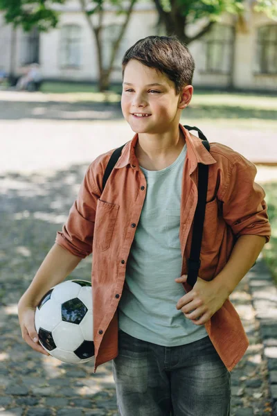Cheerful schoolboy looking away while walking in park and holding soccer ball — Stock Photo