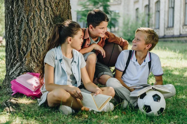 Three cheerful schoolkids talking while sitting on lawn under tree — Stock Photo