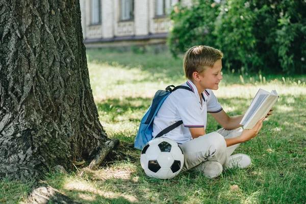 Cute smiling schoolboy reading book while sitting on lawn near soccer ball — Stock Photo