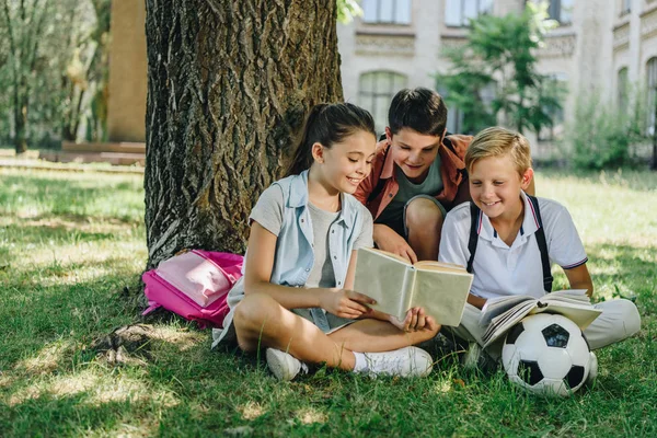 Trois camarades de classe mignons lisant le livre et souriant tout en étant assis sur la pelouse sous l'arbre — Photo de stock