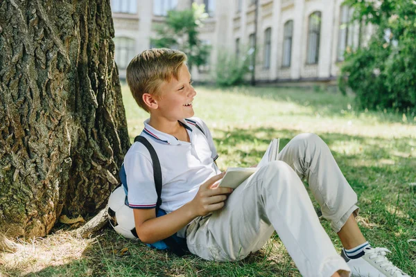 Adorable sonriente colegial sentado en el césped bajo el árbol y sosteniendo libro - foto de stock