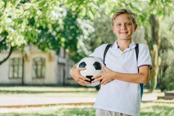 Cute cheerful schoolboy holding soccer ball and smiling at camera — Stock Photo