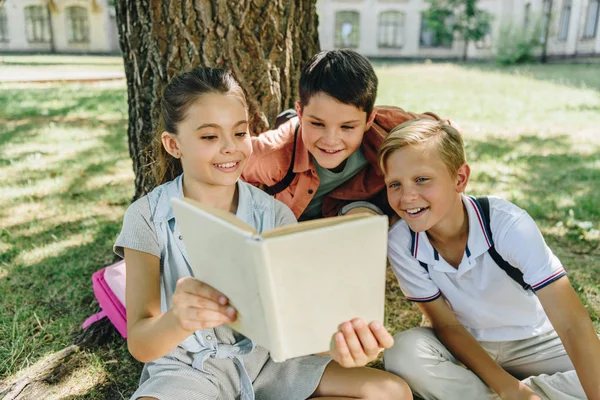 Tres escolares alegres leyendo libro mientras están sentados bajo el árbol en el parque - foto de stock