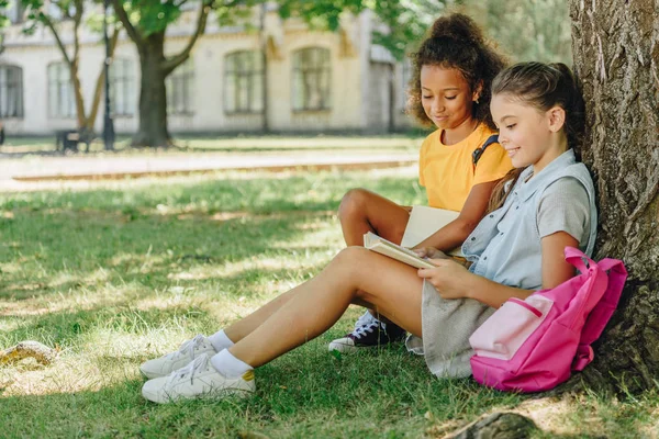 Due simpatiche studentesse multiculturali sedute sul prato sotto l'albero e leggendo insieme il libro — Foto stock