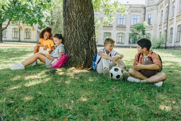 Quattro simpatici scolari multiculturali seduti su prato e albero, che parlano e leggono libri — Foto stock