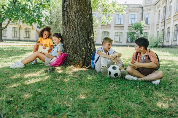 Cuatro lindos escolares multiculturales sentados en el césped y el árbol, hablando y leyendo libros - foto de stock