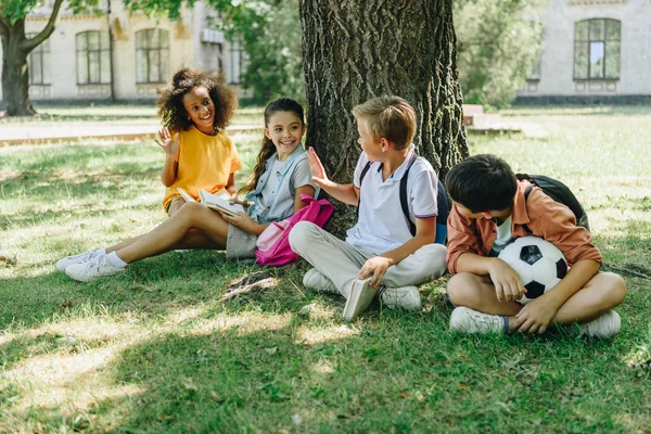 Four cute multicultural schoolkids talking while sitting on lawn under tree — Stock Photo