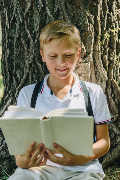 Adorable sonriente colegial sentado cerca de árbol y libro de lectura - foto de stock