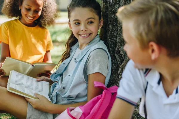 Selective focus of three cheerful multicultural classmates speaking while sitting under tree — Stock Photo