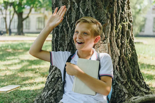 Cheerful schoolboy with book waving hand while sitting under tree and looking away — Stock Photo