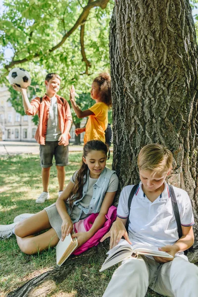 Adorables escolares sentados bajo el árbol y leyendo libros cerca de amigos multiculturales - foto de stock