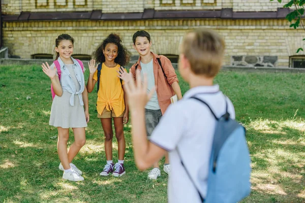 Back view of shoolboy waving hand to smiling multicultural friends — Stock Photo