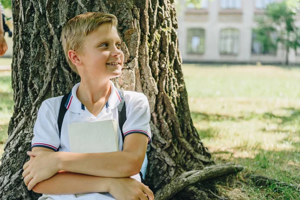 Очаровательный школьник сидит на лужайке под деревом, улыбается и смотрит в сторону — стоковое фото