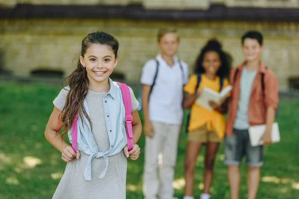 Selektiver Fokus der entzückenden Schülerin, die in die Kamera lächelt, während sie neben multiethnischen Freunden steht — Stockfoto