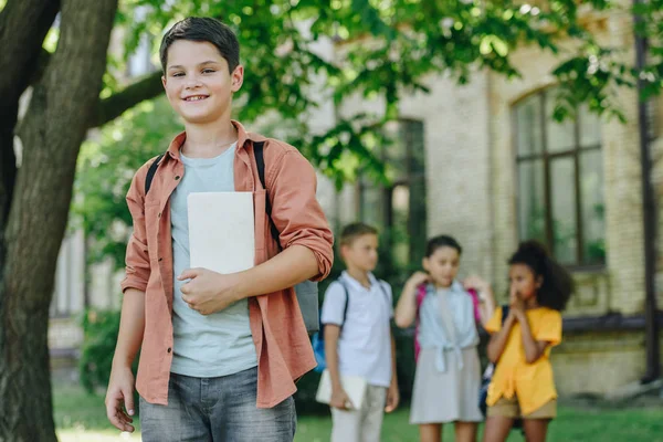Selective focus of adorable schoolboy with book smiling at camera while standing near multicultural friends — Stock Photo