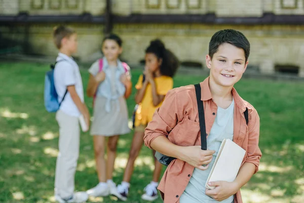 Selektiver Fokus eines lächelnden Schuljungen mit Buch, der in die Kamera schaut, während er neben multikulturellen Freunden steht — Stockfoto