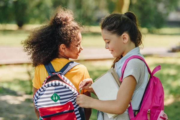 Милая школьница открывает рюкзак улыбающейся африканской американской подруги — стоковое фото