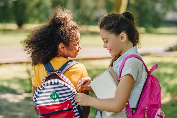 Cute schoolgirl opening backpack of smiling african american friend — Stock Photo