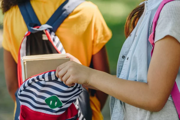 Recortado vista de colegiala embalaje libros en mochila de africano americano amigo - foto de stock