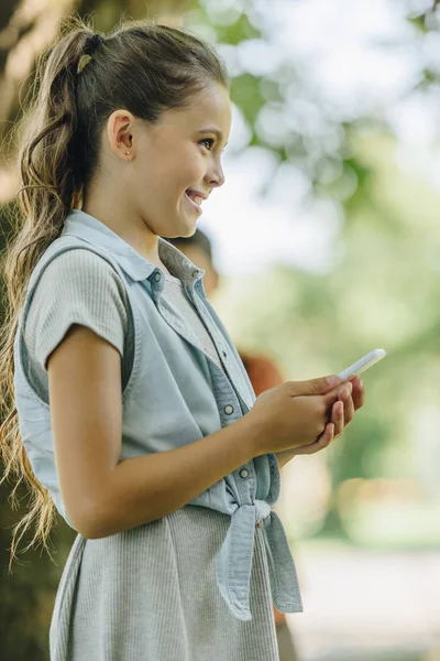Милий, усміхнений школярка посміхається, дивлячись далеко під час використання смартфона в парку — Stock Photo