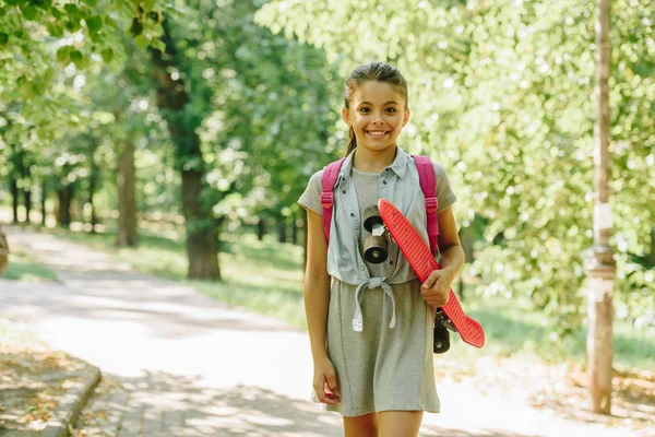 Adorable schoolgirl holding skateboard and smiling at camera in park — Stock Photo