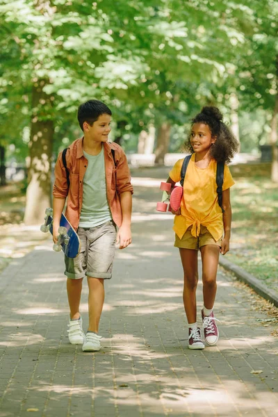 Two multicultural schoolkids walking in park while holding skateboards — Stock Photo