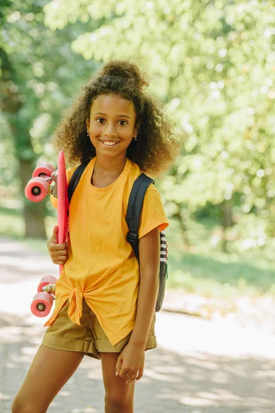 Adorable afro-américaine écolière souriant à la caméra tout en tenant skateboard — Photo de stock