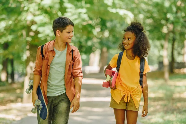 Двое улыбающихся мультикультурных школьников, гуляющих в парке, держа скейтборды — стоковое фото