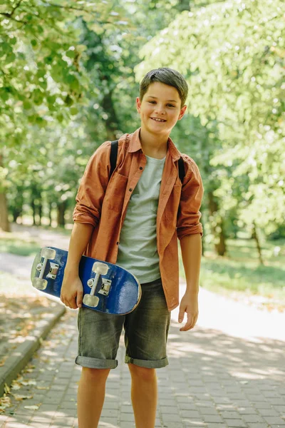 Adorable, sonriente colegial sosteniendo monopatín y mirando a la cámara en el parque - foto de stock