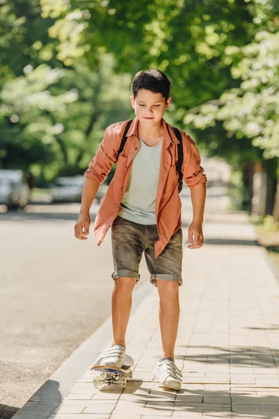 Cute schoolboy with backpack riding skateboard on sunny street — Stock Photo
