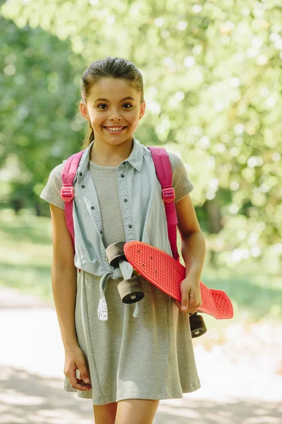 Mignonne, écolière souriante tenant skateboard et regardant la caméra dans le parc — Photo de stock