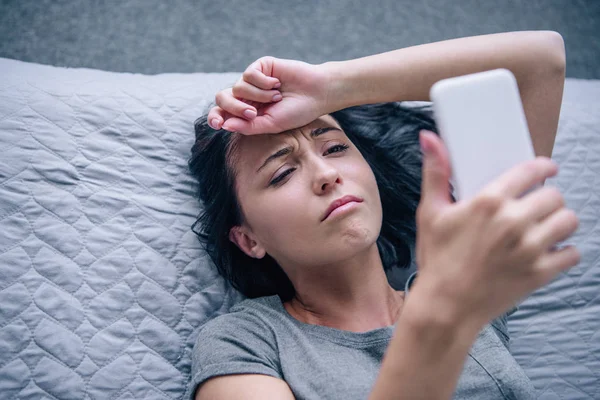 Hermosa mujer deprimida en la cama usando teléfono inteligente en el dormitorio - foto de stock